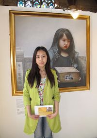 二零一三年六月，于铭慧手里拿着营救父亲的明信片站在“真善忍国际美展”画作“孤儿泪”前。