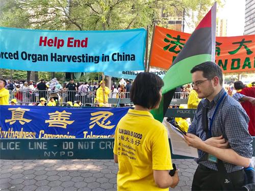 '图1：福克斯新闻记者亚当·萧（Adam Shaw，右）站在“请帮助结束发生在中国的大屠杀”（Help End Genocide in China）条幅前，采访法轮功学员简（Jane）。'
