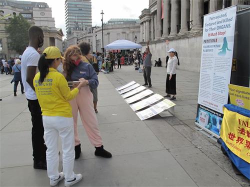'图8：二零二零年八月二十二日，在特拉法加广场，第一次看到法轮功真相的两位伦敦学生斯卡特（Scalett，正在签名的女青年）和万德瑞（Vandre，正定睛观看法轮功学员炼功男青年）签名制止迫害。'